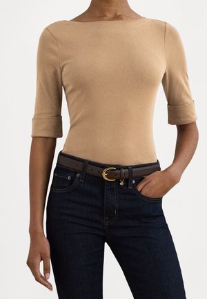 Mujer con una camiseta beige ajustada con mangas hasta el codo y vaqueros azul oscuro, complementada con un cinturón marrón con hebilla dorada.