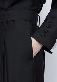 Svart skräddarsydd jacka med bälte i midjan, som har en sidoficka och slät textur. Ärmen har fem runda knappar klädda med tyg.