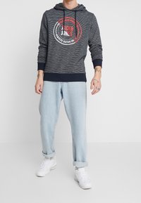 Felpa a strisce blu navy con cappuccio regolabile e design grafico. Abbinata a jeans azzurri arrotolati e sneakers bianche.