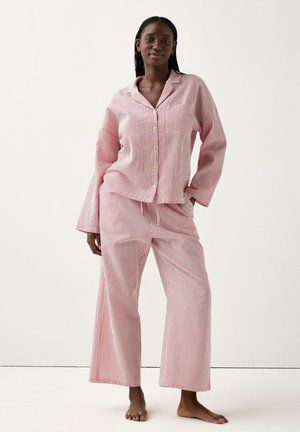 Sort kvinde stående barfodet iført et løst pink og hvidt pyjamasæt med lodrette striber og bredbenede bukser.