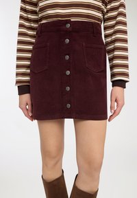 Donna che indossa una gonna burgundy in velluto a coste con bottoni sul davanti e tasche sul davanti, una maglia a righe marroni a maniche lunghe e stivali marroni.