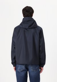 Marineblauwe waterdichte jas met een capuchon, elastische manchetten en een relaxte pasvorm. De stof lijkt glad met minimale naden en een eenvoudig ontwerp.