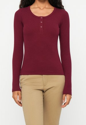 Kobieta ubrana w dopasowany bordowy top henley z długim rękawem i trzema guzikami, zestawiony z beżowymi spodniami na białym tle.