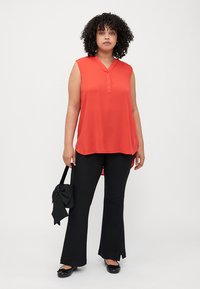 Blouse sans manches rouge avec patte de boutonnage, associée à un pantalon noir évasé. Tissu lisse et léger avec un ourlet arrière légèrement plus long.