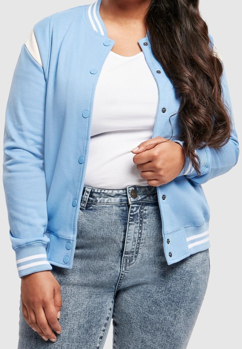 Femme portant une veste varsity bleu clair par-dessus une chemise blanche et un jean taille haute délavé à l'acide, tenant le revers de la veste avec une main.