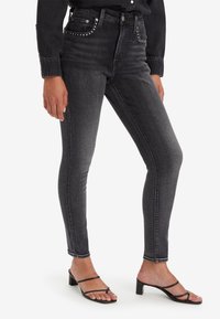 Vaqueros skinny negros de cintura alta, con tachuelas plateadas en los bolsillos delanteros y una textura ligeramente desgastada. Combinados con sandalias de tacón de tiras negras.