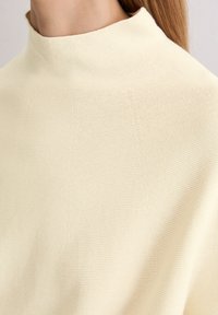 Zbliżenie osoby w kremowym, prążkowanym swetrze z wysokim kołnierzem, z długimi włosami widocznymi z jednej strony.