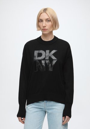 Blond person iført en sort DKNY sweater og lyseblå jeans, stående mod en ensfarvet hvid baggrund.