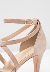 Sandal con tacco alto in suede beige, con cinturini incrociati e fibbia in tonalità oro. Texture liscia, punta affusolata e tacco sottile.