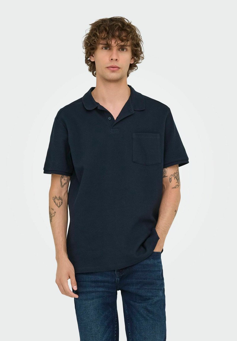 Navy polo shirt made of textured fabric, featuring a collar, short sleeves, and a chest pocket, paired with blue jeans.