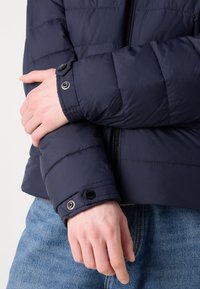 Jūros mėlynumo pūkinė striukė su siuvinėtu paviršiumi, su užtrauktuku ir sagų akcentais rankogaliuose. Dėvima ant džinsų.