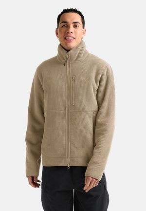 Mann trägt eine beige, gerippte Reißverschlussjacke mit hohem Kragen und schwarze Hose, lächelnd vor einem einfarbigen hellen Hintergrund.