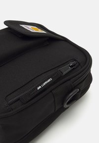 Borsa nera con una superficie tessuta e resistente, un compartimento con zip e un anello. Include una patch con un logo.