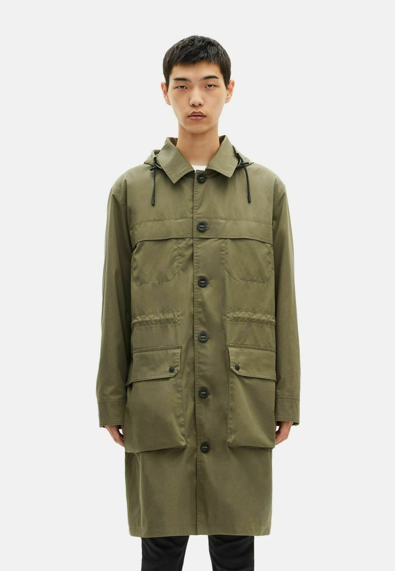 Giovane uomo che indossa un lungo cappotto verde oliva con bottoni, cappuccio e grandi tasche frontali, in piedi contro uno sfondo bianco uniforme.
