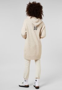 Lichtbeige gebreide hoodie-jurk met een capuchon aan de achterkant en zwarte "10" opdruk. Gecombineerd met crèmekleurige joggingbroek en witte schoenen met zwarte accenten.