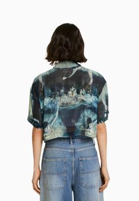 Korta tröja i genomskinligt tyg, med ett akvarelltryck med figurer, främst i blått och grönt. Korta ärmar och avslappnad passform.