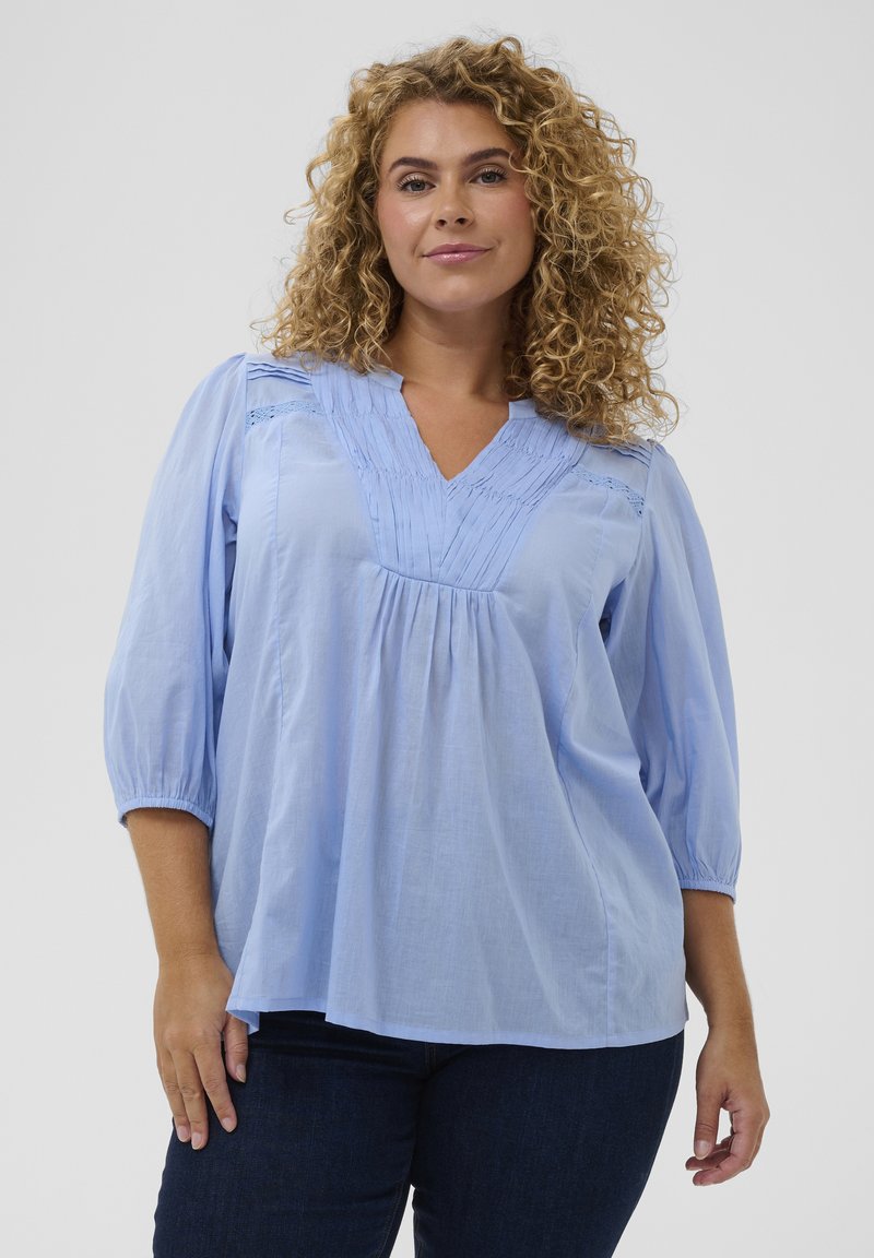 Blouse bleu clair avec un col en V, un détail plissé à l'avant et des manches trois-quarts. Confectionnée dans un tissu doux avec une coupe fluide.