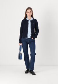 Ναυτικό πλεγμένο cardigan επάνω από ανοιχτό μπλε πουκάμισο με κολάρο, συνδυασμένο με σκούρα μπλε τζιν �και μαύρα παπούτσια. Κρατά μια μικρή μπλε τσάντα.