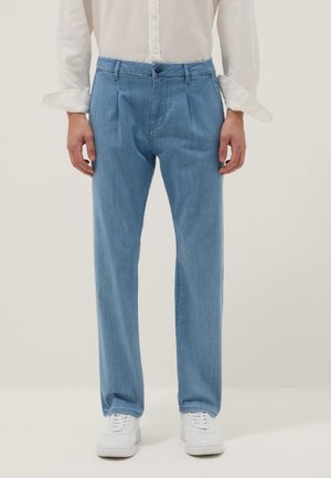 Mann trägt hellblaue plissierte Jeans, weißes Hemd mit Knöpfen und hochgekrempelten Ärmeln sowie weiße Sneakers, steht vor schlichtem Hintergrund.