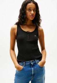 Top in cotone nero a coste con scollatura rotonda, dotato di una fila di bottoni e un piccolo logo. Indossato con jeans in denim blu a vita alta.