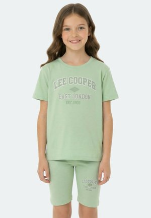 Fille souriante aux cheveux longs et ondulés portant un t-shirt vert clair Lee Cooper et un short assorti avec le texte East London.