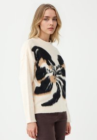 Cremfarbener Pullover mit einem großen schwarzen und beigen Schmetterlingsdesign, aus weichem Material gefertigt. Verfügt über lange Ärmel und einen runden Ausschnitt.