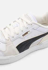 Bílé tenisky Puma s černým pruhem, béžovými semišovými detaily, bílými tkaničkami a zlatým logem Puma na boku, vyfotografované na bílém pozadí.