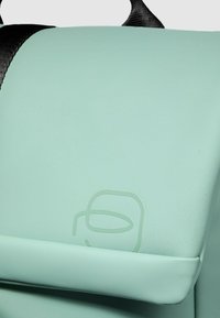 Mintgroene tas met een gladde, matte textuur, voorzien van een zwarte band. Op de voorkant is een subtiel geperst logo zichtbaar.