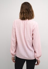 Blouse rose clair à manches longues avec un motif carré subtil, encolure froncée et poignets élastiques. Tissu lisse avec une coupe décontractée.