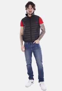 Gilet imbottito nero con colletto alto, chiusura con zip, indossato sopra una camicia rossa, abbinato a jeans blu e sneakers bianche. Stile casual.