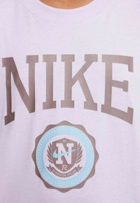 Camiseta Nike de tela rosa claro que presenta un gran texto marrón y un emblema circular azul con una "N" blanca y "Beaverton, Oregón" debajo.