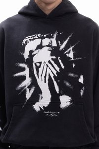 Μαύρο φούτερ με κουκούλα, διακοσμημένο με λευκό γραφικό σχέδιο που απεικονίζει πρόσωπο και χέρια. Το ύφασμα φαίνεται μαλακό· διαθέτει μπροστινή τσέπη και κουκούλα με κορδόνι.