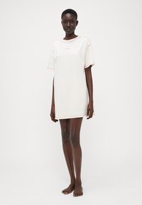 Tunika oversize a maniche corte in cotone beige chiaro con dettagli in cuciture rosa. Presenta un collo rotondo e un taglio rilassato, lunga fino al ginocchio.