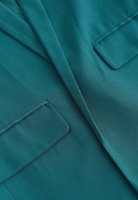 Tissu bleu sarcelle en gros plan montrant deux poches à rabat et un détail de couture, probablement partie d'un vêtement comme un blazer ou un manteau.