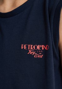 Granatowy podkoszulek bez rękawów wykonany z bawełny, z czerwoną haftowaną naszywką "PETROLIND U.S.A. Key West" z przodu. Gładka faktura.