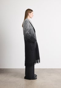 Cappotto lungo in gradiente grigio, realizzato in lana texturizzata, con orlo frangiato e tasche laterali, indossato sopra pantaloni neri e scarpe flat.