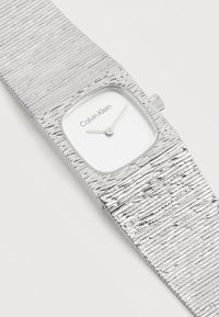 Ασημένιο ρολόι χειρός με υφή μετάλλου, τετράγωνο καντράν, δύο δείκτες και το λογότυπο Calvin Klein σε εκρού φόντο.