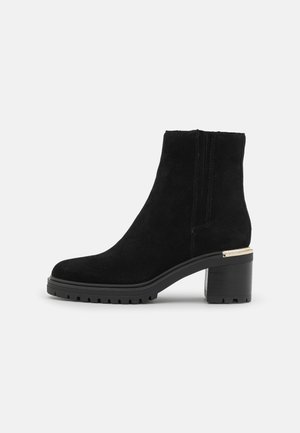 Sort ruskind ankelstøvle med blokhæl, lynlås i siden, robust sål og metalaccent øverst på hælen mod en ensfarvet hvid baggrund.