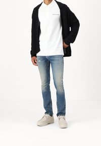 Weißes Polohemd, schwarzer Strickcardigan, hellblaue, abgenutzte Jeans und hellbeige Schuhe. Einfaches Design mit minimaler Markenkennzeichnung.