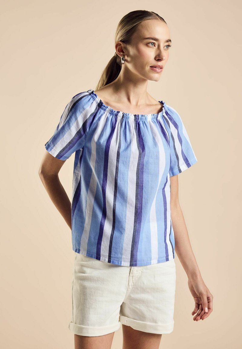 Blusa a righe blu e bianche con maniche corte e colletto arricciato, realizzata in tessuto leggero. Abbinata a pantaloni corti bianchi.