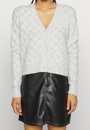 Donna che indossa un cardigan lavorato a maglia di colore grigio chiaro con bottoni e una mini gonna in pelle nera con cuciture bianche.