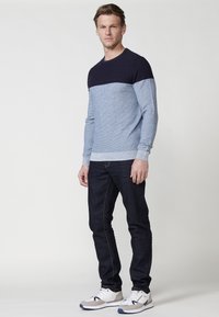 Pulover v modri in mornarski barvi s blokiranim vzorcem ter rebrastim okroglim izrezom, v kombinaciji s temno denim hlačami in belimi supergami s sivimi poudarki.
