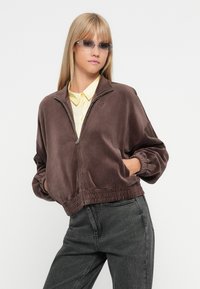 Smeđa velurska bomber jakna s patentnim zatvaračem sprijeda, elastičnim manžetama i rubom, nosi se preko svijetlo žute košulje s kragnom, u kombinaciji s tamnim trapericama.