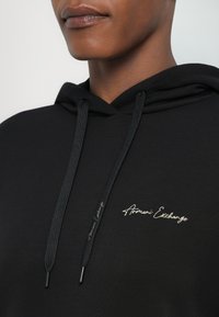 Juodas džemperis su gobtuvu ir raišteliu, su "Armani Exchange" logotipu baltu siuvinėjimu ant krūtinės. Lygus audinys su klasikinio fasono kirpimu.