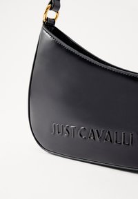 Μαύρη δερμάτινη τσάντα ώμου με λεία υφή, στρογγυλή μορφή και ανάγλυφο λογότυπο "JUST CAVALLI" σε έντονα γράμματα. Χρυσές λεπτομέρειες.