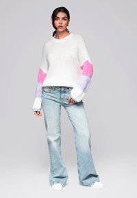 Pull en tricot blanc avec des accents rayés rose et bleu sur les manches, associé à un jean évasé délavé et des baskets blanches.