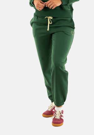 Pantalones de chándal verdes con cintura elástica y cordón dorado, que presentan un pequeño diseño de corazón. Combinados con zapatillas burdeos y calcetines blancos.