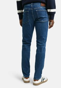 Vaqueros de denim azul de corte slim con un lavado medio, que presentan bolsillos traseros y una etiqueta de cuero en la cinturilla. Llevados con zapatillas blancas y marrones.