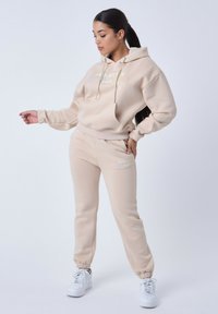 Beige hoodie en bijpassende joggingsbroek van zacht materiaal. Voorzien van een trekkoordcapuchon, geribde manchetten en geprinte logo-details.