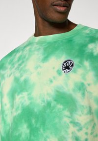 Πράσινο βαμβακερό t-shirt με τεχνική βαφής tie-dye, διαθέτει στρογγυλή λαιμόκοψη και λεπτομέρεια με μαύρο λογότυπο κρανίου. Μαλακή υφή με αποχρώσεις σε διάφορα τόνους πράσινου.